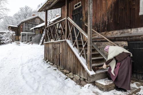 A woman dressed in historical attire sweeps the snowy stairs of an old wooden house at Luostarinmäki Museum Quarter.